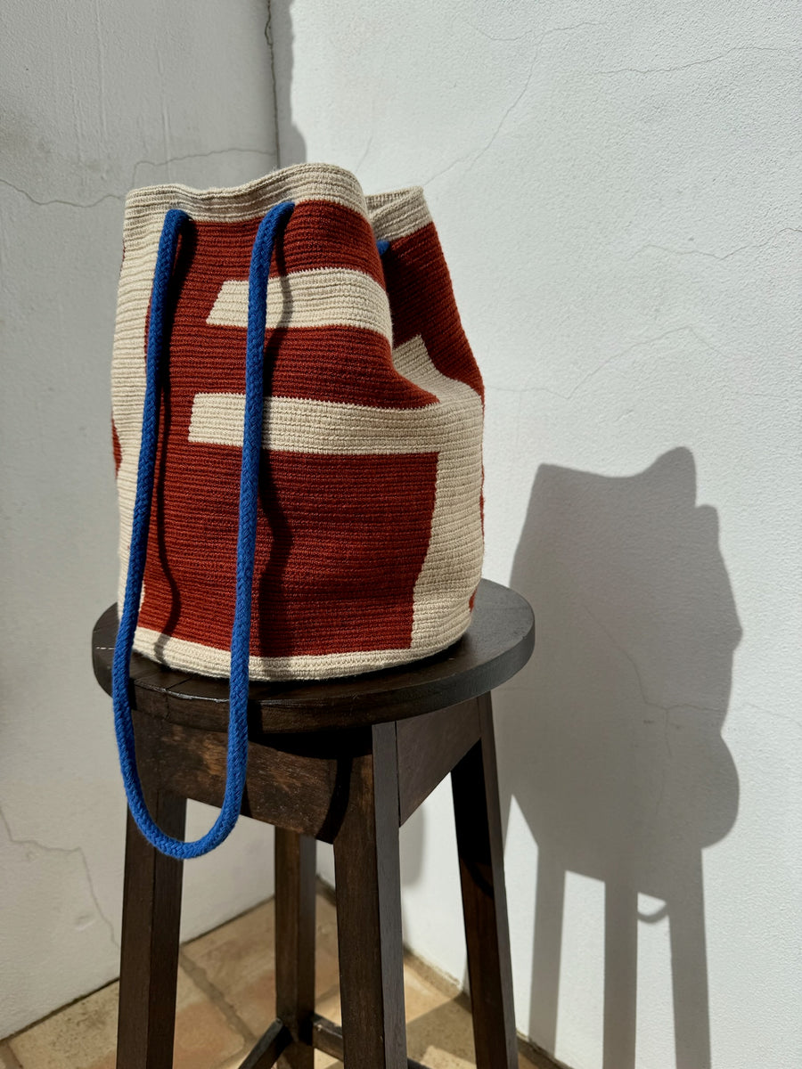 A red and beige coloured knitted cotton bag with blue straps on a wooden stool in front of a white wall.